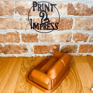 May include: A brown, miniature sofa-shaped tray. The tray is on a wooden surface, with a brick wall in the background. A black metal sign with the text "PRINT 2 IMPRESS" is above the tray.