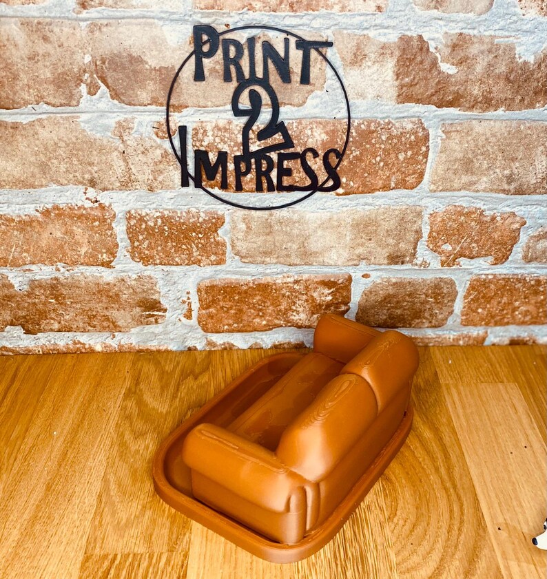 May include: A brown, miniature sofa-shaped tray. The tray is on a wooden surface, with a brick wall in the background. A black metal sign with the text "PRINT 2 IMPRESS" is above the tray.