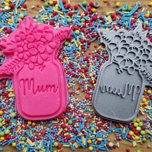 May include: Two cookie cutters in the shape of a jar with a floral design. One is pink, the other grey. The word "Mum" is inscribed on the front of each jar. The cutters are surrounded by colourful sprinkles.
