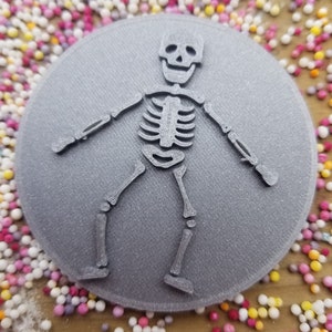 May include: A gray 3D printed cookie cutter shaped like a skeleton with a smiling face. The cookie cutter is on a background of colorful sprinkles.
