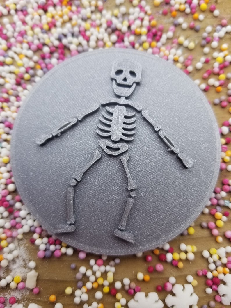 May include: A gray 3D printed cookie cutter shaped like a skeleton with a smiling face. The cookie cutter is on a background of colorful sprinkles.