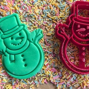 Op de afbeelding: Een groene sneeuwpopkoekjesvorm en een rode sneeuwpopkoekjesvorm liggen op een houten oppervlak bedekt met kleurrijke hagelslag. De groene vorm is gevuld met een sneeuwpopkoekje. De rode vorm is leeg.