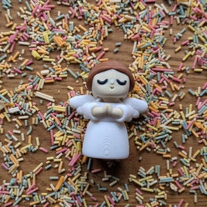 May include: A white angel figurine with brown hair and closed eyes, standing with hands clasped in prayer. The figurine is surrounded by colorful sprinkles on a wooden surface.