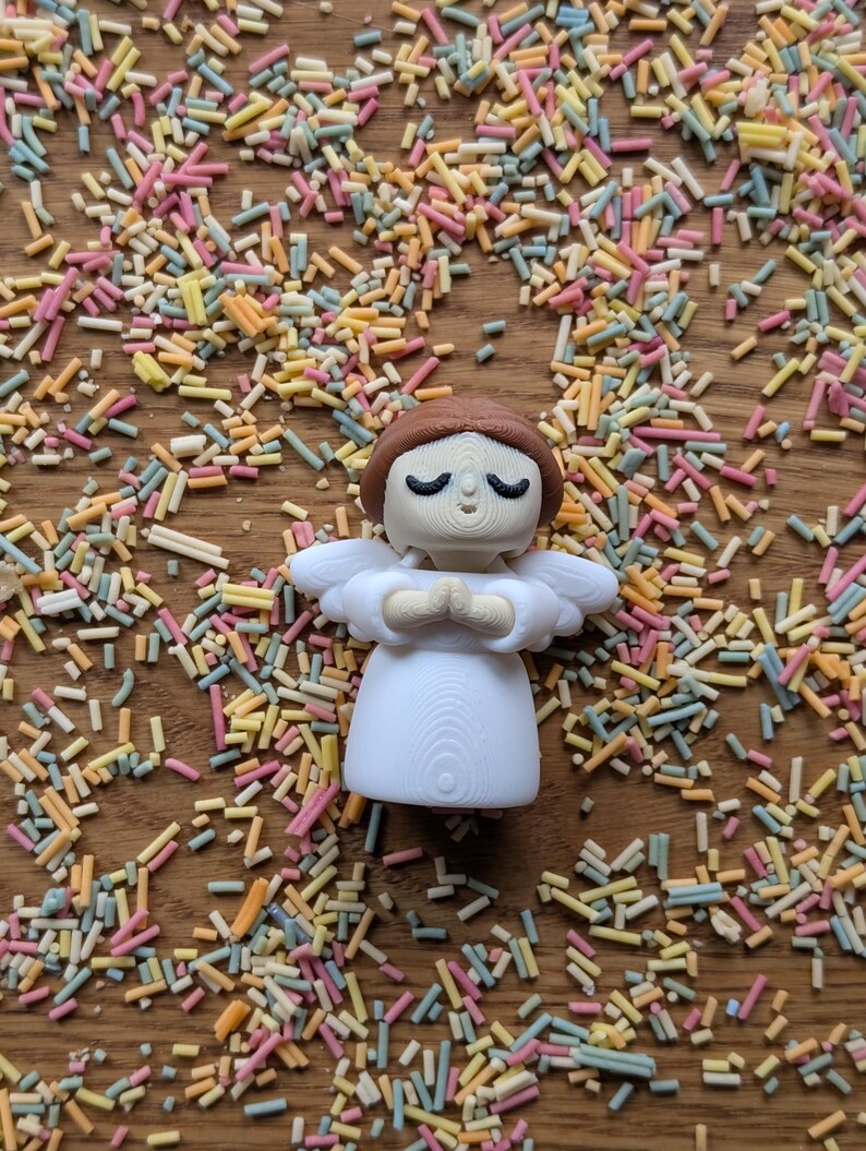 May include: A white angel figurine with brown hair and closed eyes, standing with hands clasped in prayer. The figurine is surrounded by colorful sprinkles on a wooden surface.