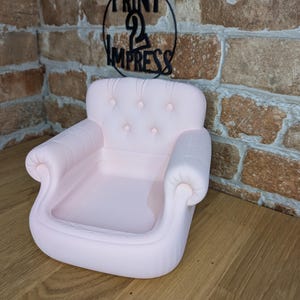 May include: A light pink miniature sofa with a tufted back and rolled arms. The small couch is a decorative item, possibly for display or as a novelty piece. The background includes a brick wall and a sign that says "PRINT 2 IMPRESS".