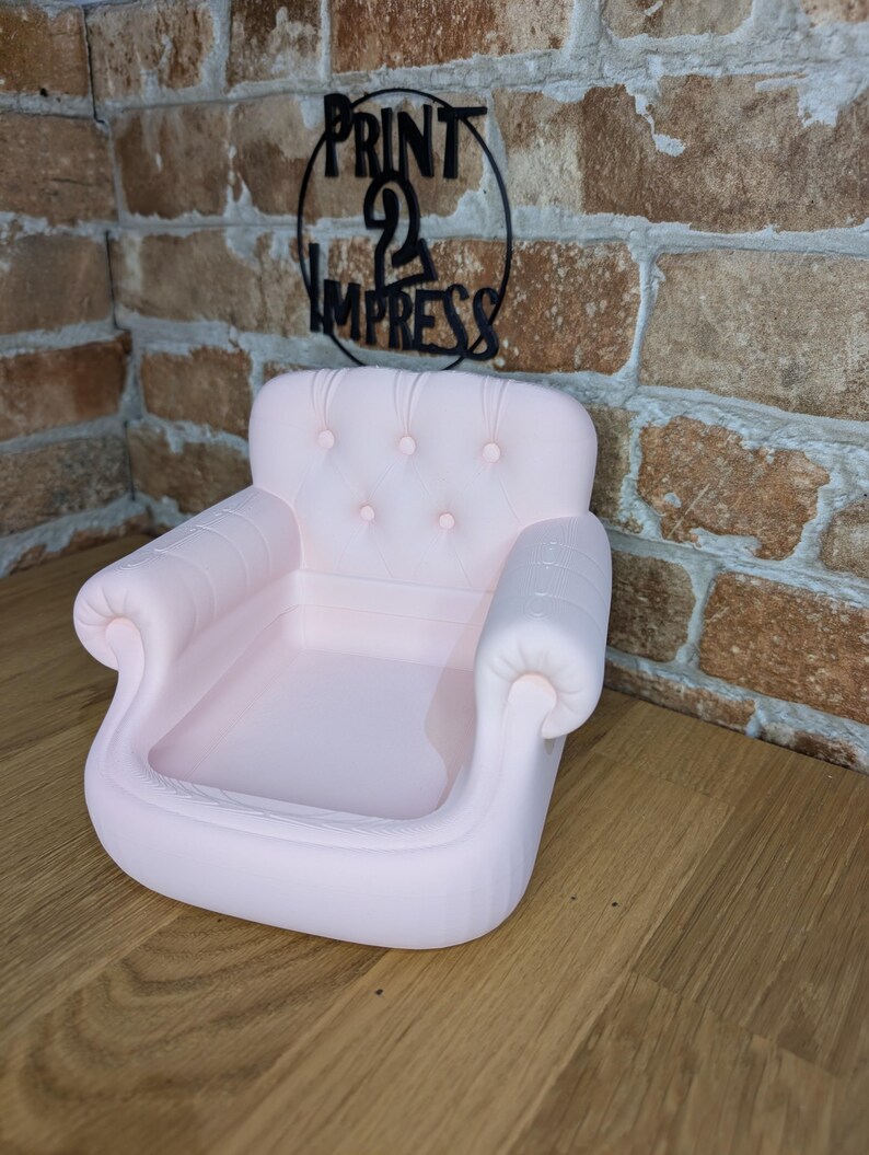 May include: A light pink miniature sofa with a tufted back and rolled arms. The small couch is a decorative item, possibly for display or as a novelty piece. The background includes a brick wall and a sign that says "PRINT 2 IMPRESS".