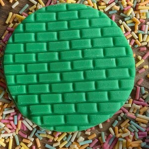 May include: A green, circular fondant decoration with a brick pattern, surrounded by colorful sprinkles. The sprinkles are in shades of pink, yellow, orange, and blue. This image is suitable for cake decorating.