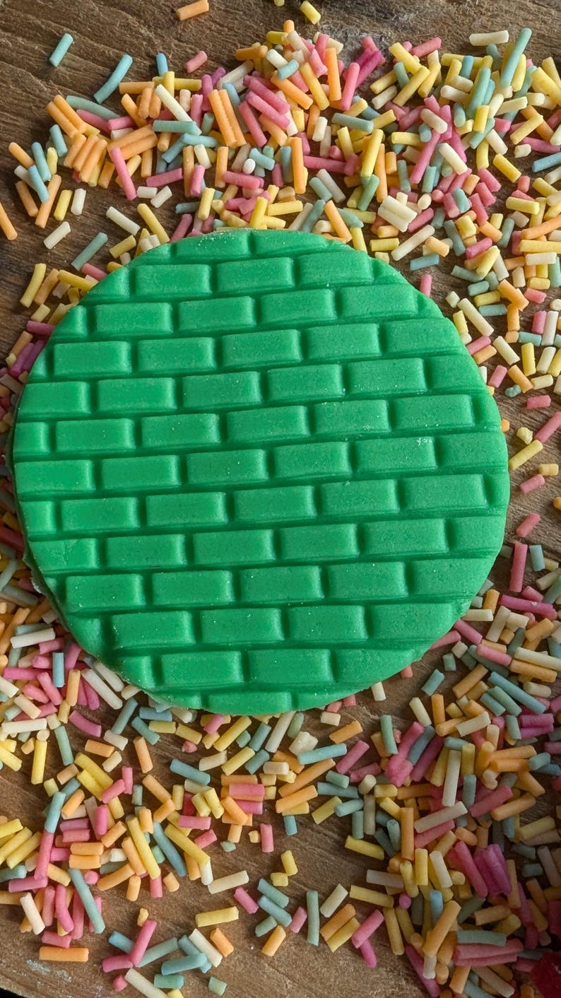 May include: A green, circular fondant decoration with a brick pattern, surrounded by colorful sprinkles. The sprinkles are in shades of pink, yellow, orange, and blue. This image is suitable for cake decorating.