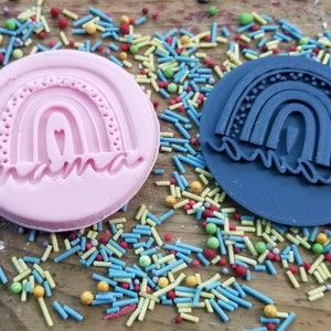 May include: Two round cookie stamps, one pink and one blue, each featuring a rainbow design with the word "mama". The stamps are surrounded by colorful sprinkles, suggesting a baking theme. The pink stamp is raised, while the blue stamp is recessed.