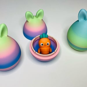 May include: Four pastel colored Easter eggs with bunny ears. One egg is open and contains a small orange carrot-shaped toy with a smiling face.