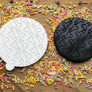 May include: Two round cookie stamps, one white and one black, each featuring a repeating pattern of a phallic symbol. The stamps are surrounded by colorful sprinkles on a wooden surface. The white stamp has a small handle.