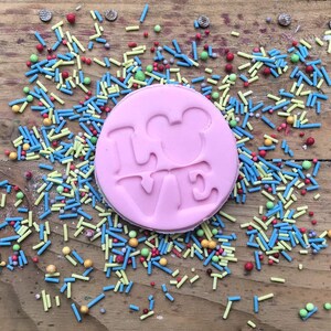 May include: A pink, circular biscuit with the word "LOVE" and a Mickey Mouse head silhouette embossed. It's surrounded by colourful sprinkles on a wooden surface.