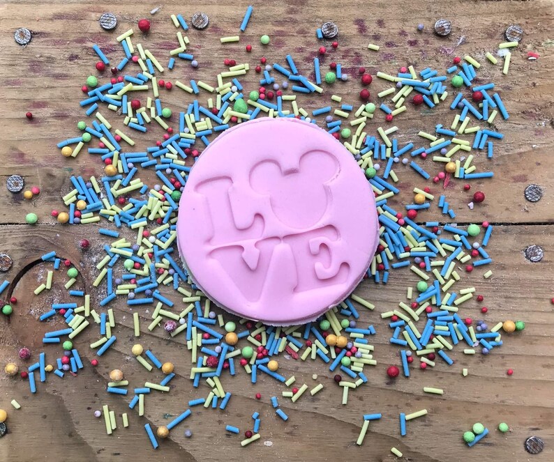May include: A pink, circular biscuit with the word "LOVE" and a Mickey Mouse head silhouette embossed. It's surrounded by colourful sprinkles on a wooden surface.