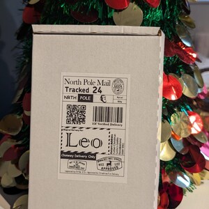 May include: A white cardboard box with a North Pole Mail label, addressed to "Leo." The label includes a QR code, barcode, and the text "Chimney Delivery Only." A festive Christmas tree with red and gold ornaments is in the background.