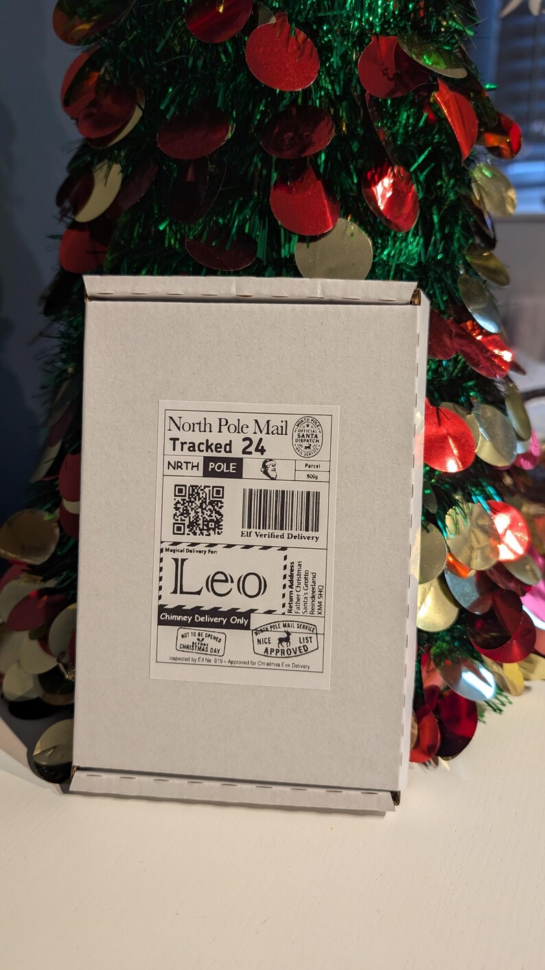 May include: A white cardboard box with a North Pole Mail label, addressed to "Leo." The label includes a QR code, barcode, and the text "Chimney Delivery Only." A festive Christmas tree with red and gold ornaments is in the background.