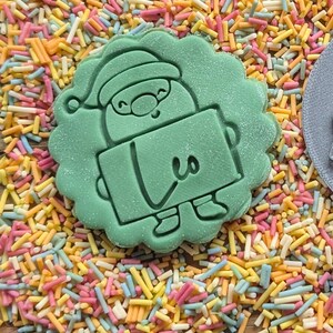 Op de afbeelding: Lichtgroene koekjesvorm in de vorm van een kerstman die een bord met het nummer 3 vasthoudt. De koekjesvorm is omgeven door kleurrijke hagelslag in roze, geel, oranje en blauw.