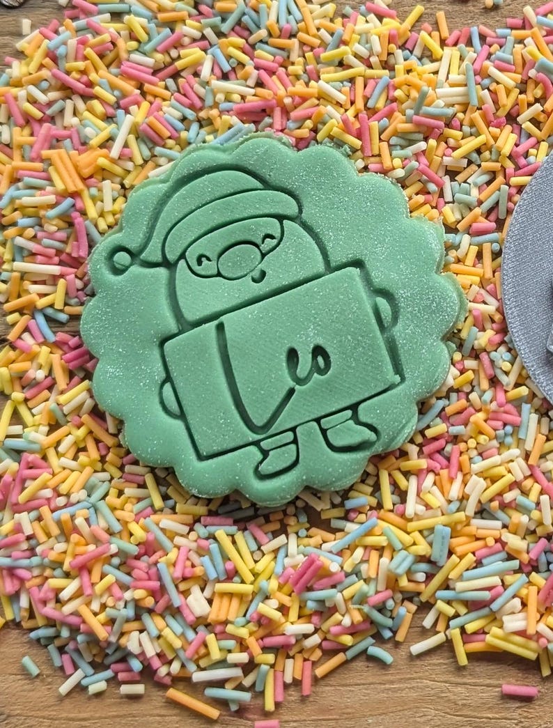 Op de afbeelding: Lichtgroene koekjesvorm in de vorm van een kerstman die een bord met het nummer 3 vasthoudt. De koekjesvorm is omgeven door kleurrijke hagelslag in roze, geel, oranje en blauw.