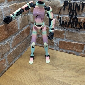 May include: A multi-jointed, articulated figure in pastel colors of pink, green, and black. The figure is posed with one arm extended. The figure is standing on a wooden surface in front of a brick wall. The text "PRINT 2 IMPRESS" is visible.
