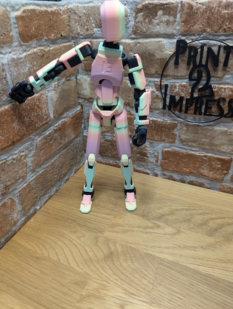 May include: A multi-jointed, articulated figure in pastel colors of pink, green, and black. The figure is posed with one arm extended. The figure is standing on a wooden surface in front of a brick wall. The text "PRINT 2 IMPRESS" is visible.