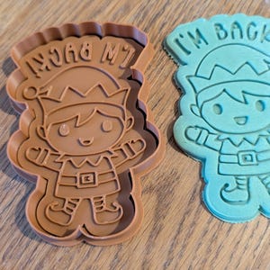 May include: Two elf-shaped cookie cutters. One is brown with the text "I'M BACK!" and the other is blue with the same text. The cutters have a scalloped edge and are designed with an elf wearing a hat and belt.