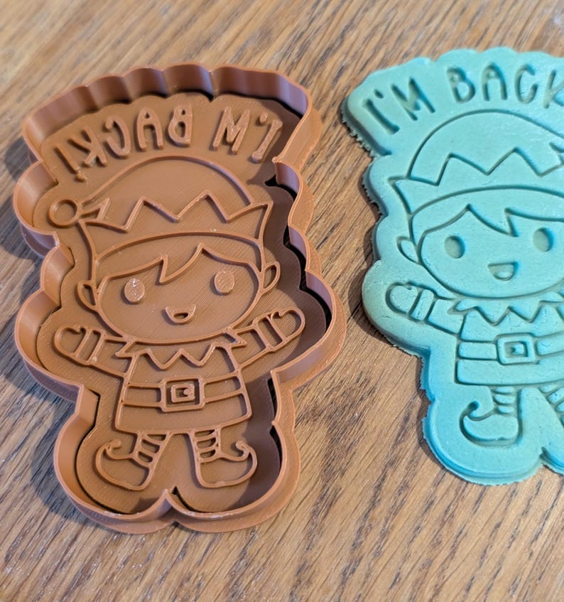 May include: Two elf-shaped cookie cutters. One is brown with the text "I'M BACK!" and the other is blue with the same text. The cutters have a scalloped edge and are designed with an elf wearing a hat and belt.