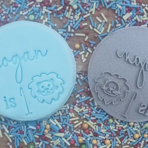 Puede incluir: Dos adornos redondos para pastel, celeste y gris, con los nombres "Logan" y "Nypal" en escritura cursiva. Cada uno tiene una cara de le&oacute;n y el n&uacute;mero "1" grabado. Rodeados de grageas coloridas.