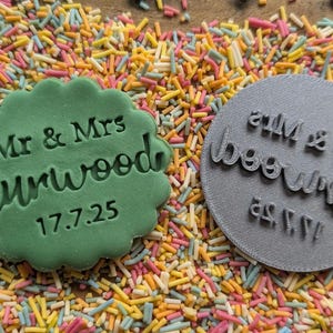 Op de afbeelding: Een groene koekje met geschulpte rand met de tekst "Mr & Mrs Curwood 17.7.25" naast een grijze ronde stempel met dezelfde tekst. De items zijn omgeven door kleurrijke hagelslag.