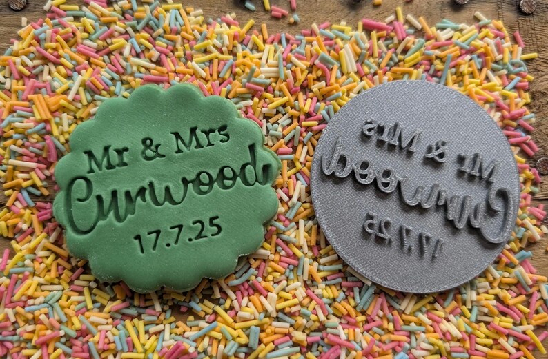 Op de afbeelding: Een groene koekje met geschulpte rand met de tekst "Mr & Mrs Curwood 17.7.25" naast een grijze ronde stempel met dezelfde tekst. De items zijn omgeven door kleurrijke hagelslag.