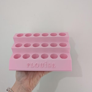 May include: A pink tiered makeup organizer with multiple circular openings. The word "PLouise" is printed on the front. The organizer is held up against a white wall.