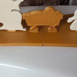 May include: A gold-colored nativity scene with glitter accents. The scene includes figures in white and a gold manger. The base is a gold color. The figures are arranged on a gold-colored base.