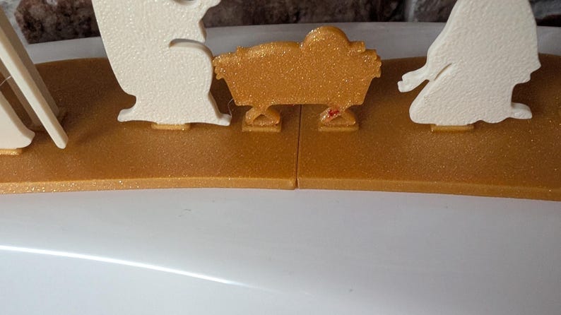 May include: A gold-colored nativity scene with glitter accents. The scene includes figures in white and a gold manger. The base is a gold color. The figures are arranged on a gold-colored base.