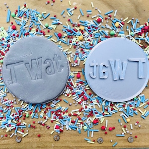 Op de afbeelding: Twee grijze, ronde koekjesstempels, &eacute;&eacute;n met het woord "TWAT" erop gedrukt en de andere met het woord in spiegelbeeld. De stempels zijn omgeven door kleurrijke hagelslag op een houten oppervlak.