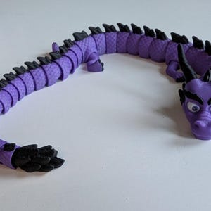K&ouml;nnte beinhalten: Ein lila-schwarzes, segmentiertes Drachenspielzeug mit einem detaillierten Kopf und gro&szlig;en Augen. Der Drache ist in einer geschwungenen Form aufgestellt.