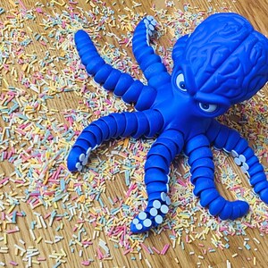 May include: A bright blue octopus toy with a detailed brain-like head and expressive eyes. The octopus is surrounded by colorful sprinkles on a wooden surface, creating a playful and whimsical scene.