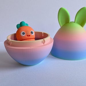May include: A pastel pink, blue, and green egg-shaped container with a removable lid. The lid has two green bunny ears. Inside the container is a small orange carrot-shaped toy with a green leafy top and a smiling face.