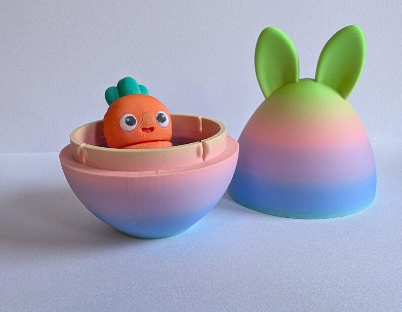 May include: A pastel pink, blue, and green egg-shaped container with a removable lid. The lid has two green bunny ears. Inside the container is a small orange carrot-shaped toy with a green leafy top and a smiling face.