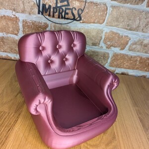May include: A small, mauve-colored miniature armchair with a button-tufted back and curved armrests. The chair is set against a brick wall with the text "Print 2 Impress" in a circular logo.