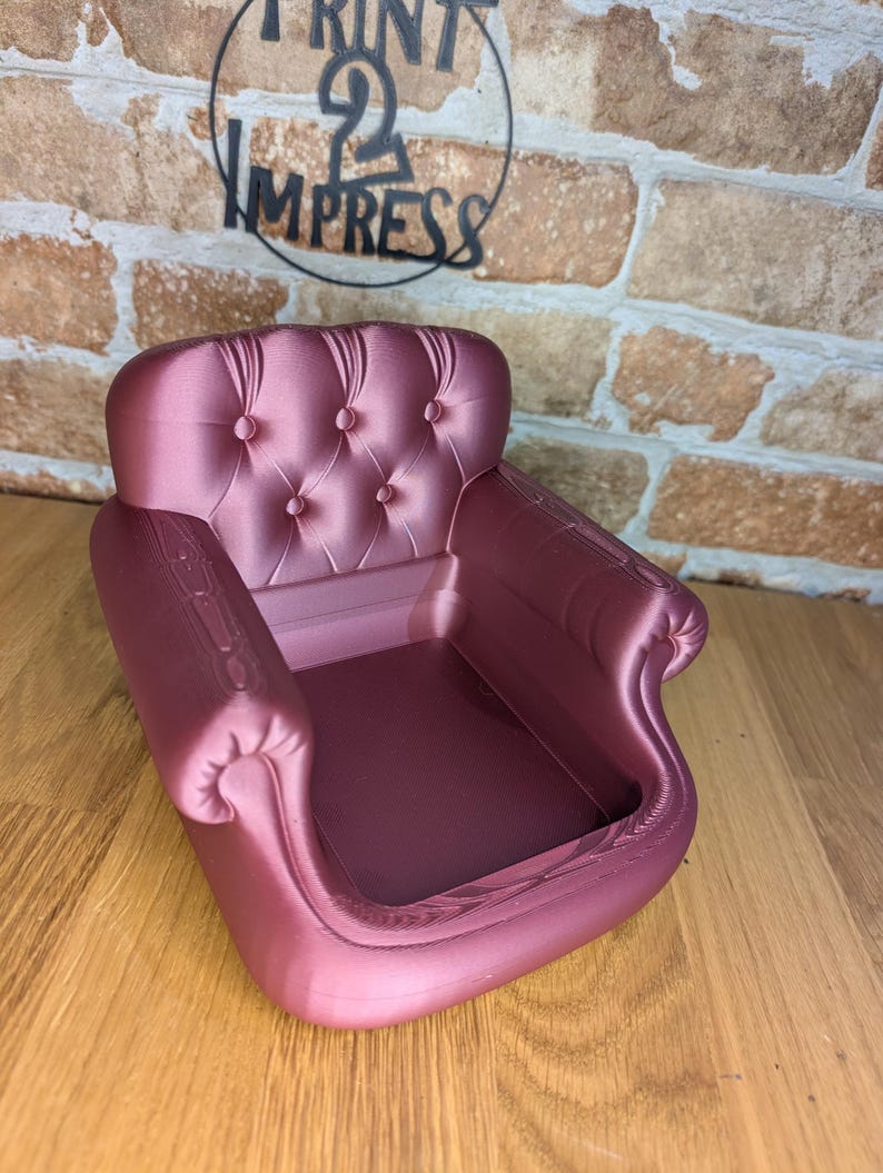 May include: A small, mauve-colored miniature armchair with a button-tufted back and curved armrests. The chair is set against a brick wall with the text "Print 2 Impress" in a circular logo.