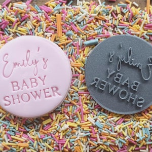 Puede incluir: Dos cortadores de galletas redondos, rosa y gris, con el texto "Emily's Baby Shower" en ellos. Los cortadores est&aacute;n sobre un fondo de chispas de colores.