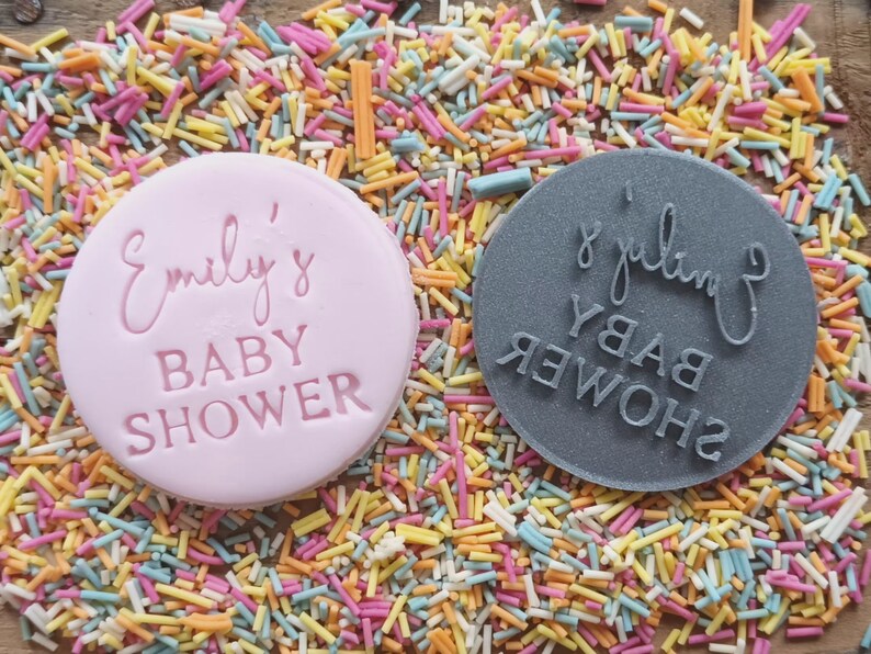Puede incluir: Dos cortadores de galletas redondos, rosa y gris, con el texto "Emily's Baby Shower" en ellos. Los cortadores est&aacute;n sobre un fondo de chispas de colores.