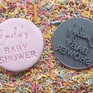 May include: Two round pink and grey cookie cutters with the text "Emily's Baby Shower" and "Emily's Baby Shower" on them. The cookie cutters are surrounded by colourful sprinkles.