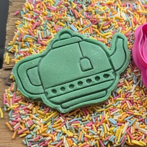 May include: A green teapot-shaped cookie cutter sits on a wooden surface covered in colorful sprinkles. The cutter has detailed features, including a spout, handle, and lid. The sprinkles are a mix of pastel colors.