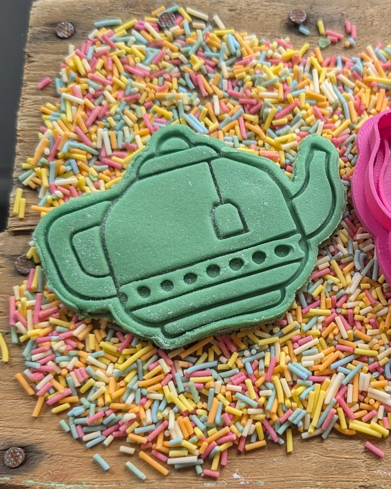 May include: A green teapot-shaped cookie cutter sits on a wooden surface covered in colorful sprinkles. The cutter has detailed features, including a spout, handle, and lid. The sprinkles are a mix of pastel colors.