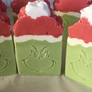 May include: Four handmade Grinch-themed soap bars. Each bar is layered with red, white, and green soap and features a Grinch face design.