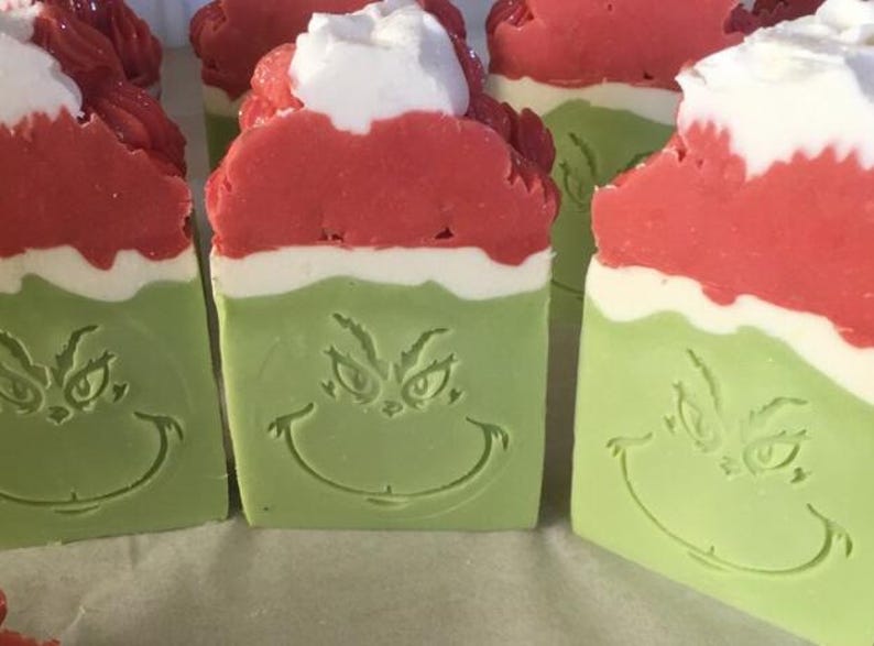 May include: Four handmade Grinch-themed soap bars. Each bar is layered with red, white, and green soap and features a Grinch face design.