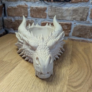 May include: A cream-colored dragon head planter with horns and detailed scales. The dragon's mouth is open, and the planter has a bowl-like shape. The word "IMPRESS" is visible in the background.