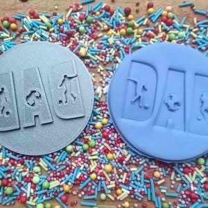 May include: Two round, gray and blue, cake decorations with the word "DAD" and soccer player figures. The decorations are surrounded by colorful sprinkles, creating a festive and playful scene.
