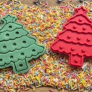 Puede incluir: Dos cortadores de galletas con forma de &aacute;rbol de Navidad, uno verde y otro rojo, sobre una cama de chispas de colores. Los cortadores tienen una estrella y puntos decorativos. Un tema festivo de vacaciones.