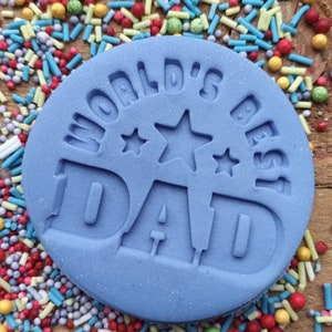 May include: A light blue, circular fondant with the words "WORLD'S BEST DAD" embossed on the surface. The fondant is surrounded by colorful sprinkles on a wooden surface. The text includes star shapes.
