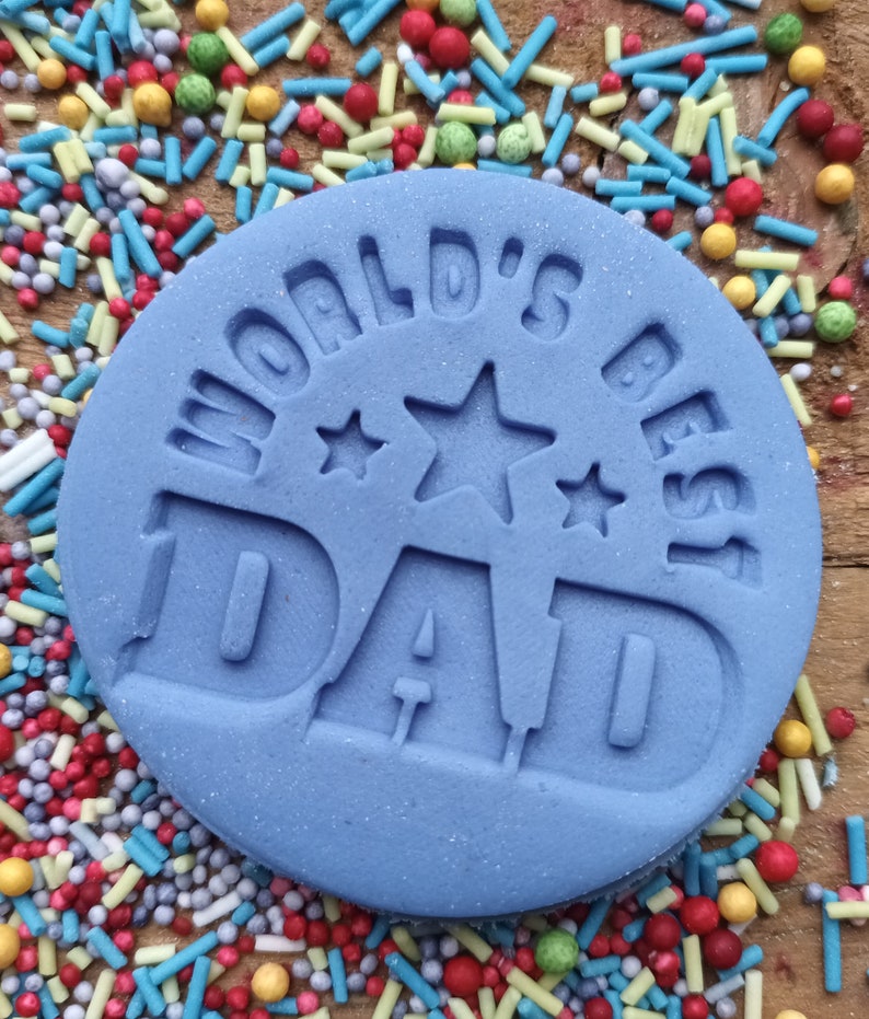 May include: A light blue, circular fondant with the words "WORLD'S BEST DAD" embossed on the surface. The fondant is surrounded by colorful sprinkles on a wooden surface. The text includes star shapes.